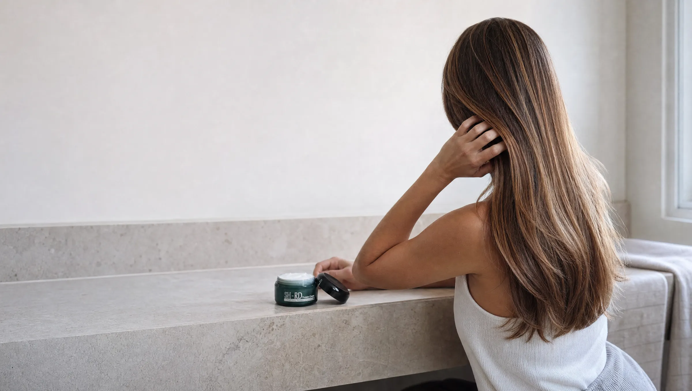 A woman runs her fingers through long, smooth, glossy hair at a bathroom vanity, an open jar of SH-RD Protein Cream on the counter